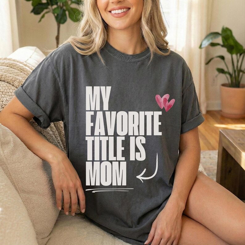 May include: Dark gray t-shirt with the text "MY FAVORITE TITLE IS MOM" in white block letters. Two pink hearts are above the word "MOM". The shirt has rolled-up sleeves.