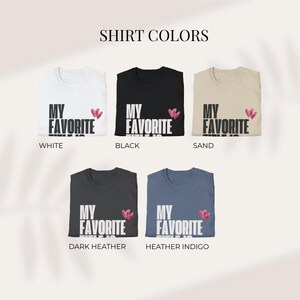 May include: A display of five t-shirts in various colors: white, black, sand, dark heather, and heather indigo. Each shirt features the text "MY FAVORITE" in white, with a small pink heart design. The text "SHIRT COLORS" is at the top.