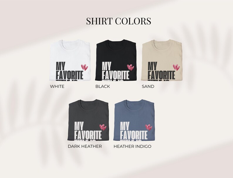 May include: A display of five t-shirts in various colors: white, black, sand, dark heather, and heather indigo. Each shirt features the text "MY FAVORITE" in white, with a small pink heart design. The text "SHIRT COLORS" is at the top.