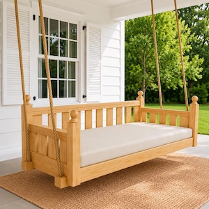 May include: A light-colored wooden porch swing bed featuring a rectangular frame with vertical slats, four corner posts topped with spherical finials, and a thick, cream-colored mattress cushion, suspended by two thick, natural-fiber ropes.