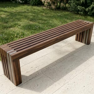 May include: A modern wooden bench with a slatted seat and rectangular legs. The bench is made of dark brown wood with silver accents and is placed outdoors on a stone patio, with green grass and foliage in the background.
