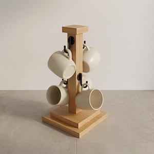 May include: A wooden mug tree with six cream-coloured mugs hanging from black hooks. The mug tree is made of light-coloured wood and has a square base and top. The mugs are evenly spaced around the central post.