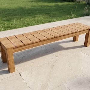 May include: A wooden bench with a slatted seat and four legs. The bench is made of light brown wood and is placed on a light-coloured stone surface, with green grass in the background. The bench is approximately 152 cm long.