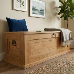 Peut inclure: Un coffre de rangement rectangulaire en bois de chêne clair. Un coussin en velours bleu marine est posé dessus. Une couverture grise et blanche à motifs et à franges est drapée sur le coffre. Une clé et une poignée en laiton sont visibles.