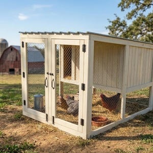 Op de afbeelding: Een crèmekleurige houten kippenhok met een metalen dak en deuren en zijkanten van gaas. Zwarte scharnieren bevestigen de deuren. Kippen zitten in het hok, dat op gras staat, met een schuur op de achtergrond.