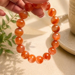 Könnte beinhalten: Ein elastisches Armband aus glatten, runden, durchscheinenden orangefarbenen und roten Karneolperlen. Die Perlen weisen natürliche Variationen in der Farbintensität und wolkenartige Innenstrukturen auf und sind auf einem elastischen Band aufgereiht.