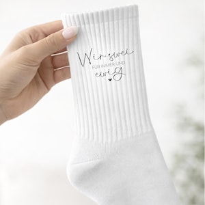 Könnte beinhalten: Weiße gerippte Socken mit dem Text "Wir zwei für immer und ewig" in schwarzer Schrift. Die Socken werden von einer Hand gehalten. Der Text bedeutet "Wir zwei für immer und ewig".