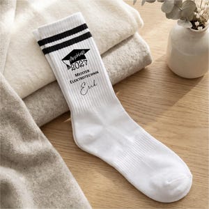 Könnte beinhalten: Ein weißer Crew-Socken mit zwei schwarzen Streifen am oberen Rand und einem schwarzen Grafikdruck. Das Design zeigt ein Doktorhut-Symbol, den Text "Abschluss 2027", "MEISTER ELEKTROTECHNIK" sowie den Namen "Erik" in einer geschwungenen Schreibschrift.