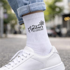 Op de afbeelding: Een witte geribbelde enkelsok met een zwarte afbeelding van een bloem en de tekst G&auml;rtner AUS LEIDENSCHAFT. De sok wordt gedragen met een blauwe spijkerbroek en een witte sneaker, met een strak ontwerp voorzien van een botanisch motief.