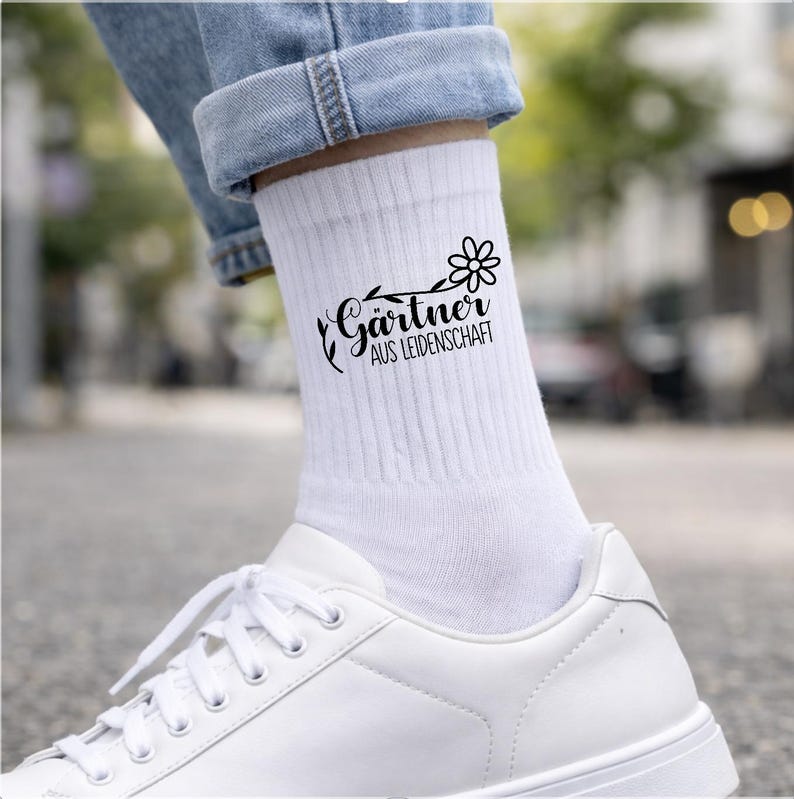 Op de afbeelding: Een witte geribbelde enkelsok met een zwarte afbeelding van een bloem en de tekst G&auml;rtner AUS LEIDENSCHAFT. De sok wordt gedragen met een blauwe spijkerbroek en een witte sneaker, met een strak ontwerp voorzien van een botanisch motief.