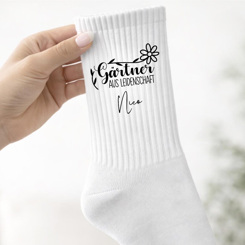 Op de afbeelding: Een witte geribbelde sok met een zwarte grafische print van een bloem, bladerige ranken en de Duitse tekst G&auml;rtner AUS LEIDENSCHAFT, gevolgd door de naam Nico in een cursief lettertype onder het bloemmotief.