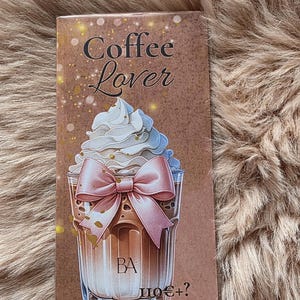 Könnte beinhalten: Eine braune Karte mit der Aufschrift "Coffee Lover" und einer Illustration eines Kaffeegetränks mit Schlagsahne und einer rosa Schleife. Das Getränk befindet sich in einem durchsichtigen Glas, mit goldenen Glitzerakzenten.