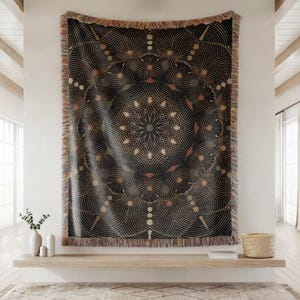 May include: Black woven throw blanket with a geometric design in gold, beige, and olive green. The blanket is rectangular with a fringed edge. The design features concentric circles and floral patterns, creating a decorative textile.