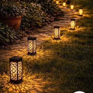 May include: A nighttime garden scene features a stone pathway illuminated by several decorative solar lanterns. The lanterns are rectangular with intricate cut-out patterns, casting warm light on the grass and path.
