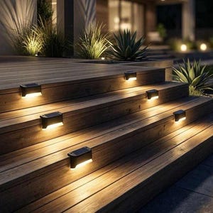 May include: Outdoor solar-powered step lights illuminate a wooden deck and staircase. The black rectangular lights emit a warm, inviting glow, enhancing the natural wood grain. Decorative plants are visible in the background, creating a welcoming outdoor space.