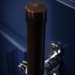 May include: A copper-toned cabinet pull with the text "Foundry Copper Collection" and "Small Format Pull - Ember Patina". The pull has a cylindrical design with a dark brown finish and silver fittings. The image is a close-up.