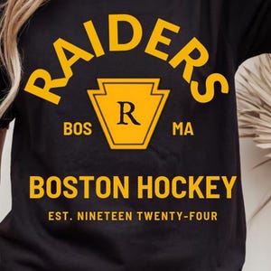 Könnte beinhalten: Schwarzes T-Shirt mit dem Wort "RAIDERS" in Gelb, das sich über einem gelben Keystone-Emblem mit einem gelben "R" wölbt. Darunter stehen "BOS" und "MA". Die Worte "BOSTON HOCKEY" sind gelb gedruckt, darunter "EST. NINETEEN TWENTY-FOUR".