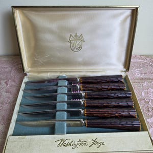 May include: A set of six steak knives with silver-toned metal blades and dark brown textured handles, presented in a rectangular box with a cream-colored interior. The box lid features a gold crest and the text Washington Forge is printed below.
