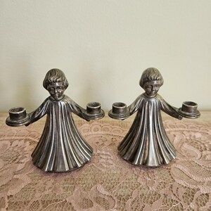 May include: Two silver candle holders in the shape of figures, each holding two candle cups. The figures have detailed faces and flowing gowns. The candle holders are displayed on a pink lace tablecloth.