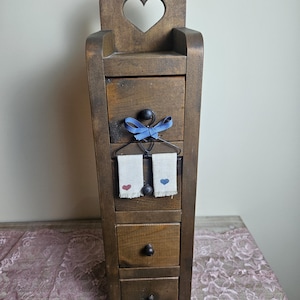 May include: A tall, dark brown wooden storage unit with four stacked drawers, each featuring a small round knob. The top section has a heart-shaped cutout. A decorative wire hanger with a blue ribbon and two small fabric towels hangs from the top drawer.