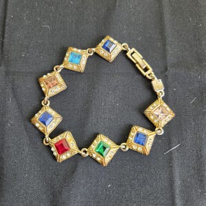 May include: A gold-tone metal link bracelet featuring eight square-shaped settings, each containing a multi-colored gemstone in shades of red, green, blue, and light brown, surrounded by small clear crystals with a fold-over clasp closure.