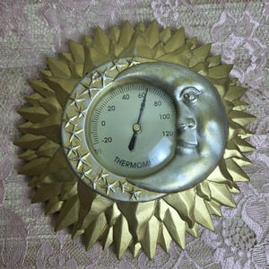 May include: A circular wall thermometer featuring a gold sunburst frame with pointed rays. A silver crescent moon with a human face is positioned on the right side. The dial displays numbers from -20 to 120 with the word THERMOMETER printed below.