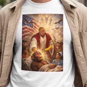 May include: White t-shirt with a parody image. The image shows a man in a red robe holding a banana, touching a chimpanzee in a bed. The background includes an American flag, angels, and other figures.