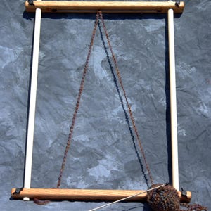 May include: A rectangular wooden frame for needlework featuring two horizontal oak bars and two vertical light-colored dowels secured with black wing nuts. A brown yarn strand hangs in a triangle shape from the top bar, with a ball of variegated brown yarn and a long metal needle resting on the bottom bar.