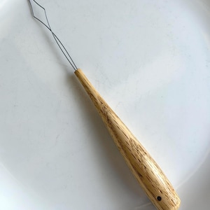 May include: A wooden needle threader with a diamond-shaped wire loop. The handle is made of light-colored wood with a textured grain. The tool is designed for threading needles and other small openings.