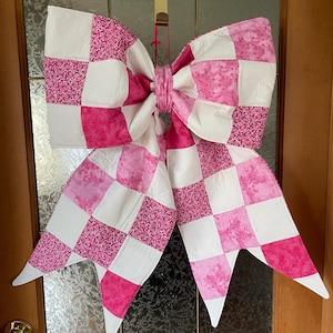 May include: A large, decorative bow made from quilted fabric, with a patchwork design of white, pink, and patterned pink squares. The bow is suspended on a door, with a red ribbon. A festive decoration.
