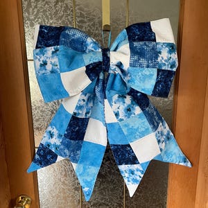 May include: A large fabric bow in various shades of blue and white, with a patchwork design. The bow has floral and geometric patterns. It is hanging on a glass door, adding a decorative touch to the entrance.
