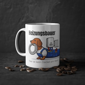 Op de afbeelding: Witte keramische mok met een cartoon teckel in blauwe overalls, die een pijp vasthoudt. De mok heeft de tekst "Heizungsbauer" en "hier ist kein Dackel am Werk". Koffiebonen zijn verspreid.