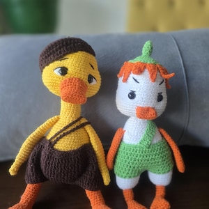 May include: Two handmade crochet duck dolls. One is yellow with brown overalls and a brown cap. The other is white with green overalls and orange hair. Both have orange beaks and feet.