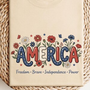 May include: Cream-coloured t-shirt featuring the word "AMERICA" in red, white, and blue, with floral and star designs. The shirt also displays the words "Freedom, Brave, Independence, Power". The t-shirt is folded on a woven, circular surface.