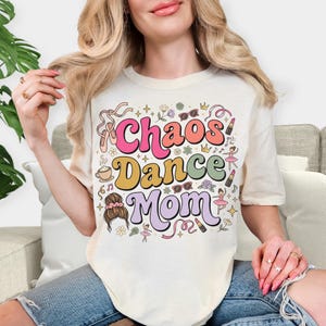 Könnte beinhalten: Cremefarbenes T-Shirt mit dem Schriftzug "Chaos Dance Mom" in farbenfrohen Retro-Lettern. Das Design umfasst Ballettschuhe, Lippenstift und Noten. Ein lässiges Kleidungsstück für den Alltag.