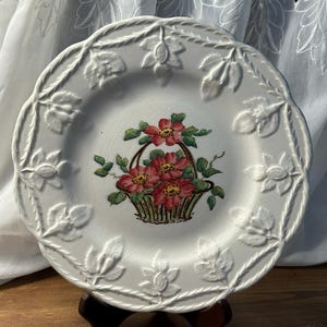 May include: White ceramic plate featuring a central floral basket design. The plate has a scalloped edge with embossed floral and leaf patterns. The basket contains red flowers and green leaves. The plate is displayed on a wooden stand.