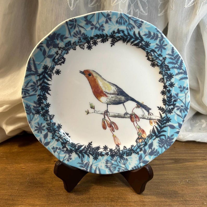 Peut inclure: Assiette d&eacute;corative en c&eacute;ramique avec une illustration d&eacute;taill&eacute;e d'un rouge-gorge perch&eacute; sur une branche avec des gousses. L'assiette a un centre blanc et une bordure florale bleue. L'assiette est pr&eacute;sent&eacute;e sur un support en bois brun fonc&eacute;.