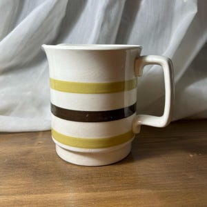 Peut inclure: Mug en céramique blanche avec des rayures horizontales vert olive et marron. La tasse a une anse carrée et un dessus légèrement évasé. La tasse est posée sur une surface en bois.