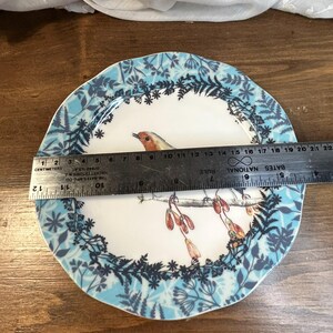 Peut inclure: Une assiette d&eacute;corative en c&eacute;ramique avec un motif d'oiseau et de fleurs. L'assiette a un centre blanc avec une illustration d'oiseau et une bordure florale bleue et blanche. L'assiette mesure environ 20 cm de diam&egrave;tre.
