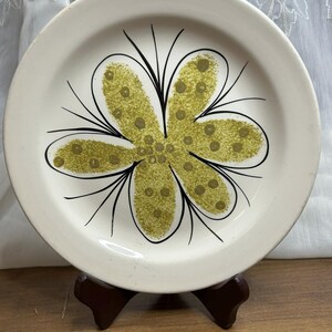 May include: A white ceramic plate featuring a large, stylised flower design in green and black. The flower has rounded petals with dotted details. The plate is displayed on a wooden stand, showcasing a vintage aesthetic.