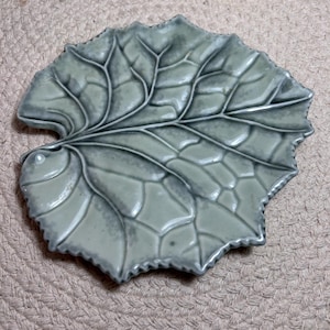 May include: A ceramic dish shaped like a large leaf with a glossy, sage green glaze. The surface features raised, detailed veins that create a textured, organic pattern across the entire piece, with scalloped edges following the leaf shape.
