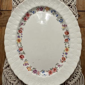 May include: An oval-shaped white ceramic serving platter featuring a scalloped edge with a textured basket-weave pattern. The centre of the platter is plain white, surrounded by a colourful floral garland border with red, yellow, purple, and blue flowers.