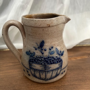 May include: A beige stoneware pitcher with a speckled texture and a single handle. The front features a blue embossed design of a basket filled with berries and leaves, with a small pouring spout at the top rim and a smooth interior finish.