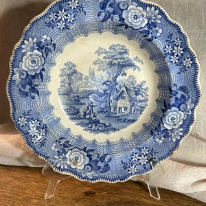 Puede incluir: Un plato de cerámica redondo con borde festoneado, que presenta un patrón de transferencia en azul y blanco. El centro muestra un paisaje rural con una cabaña, árboles y figuras, mientras que el borde exhibe un diseño floral y de red.