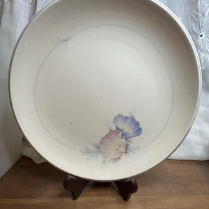 May include: A round, off-white ceramic plate with a silver rim. The plate features a delicate floral design with pink and blue flowers. A thin blue line circles the center of the plate. The plate is displayed on a brown wooden stand.