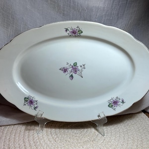 May include: An oval-shaped white ceramic serving platter featuring a delicate floral pattern of purple violets with green leaves. The design includes a central cluster of flowers and three smaller floral motifs positioned along the scalloped rim.
