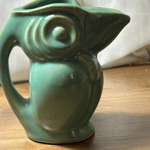 May include: A green ceramic jug shaped like a bird with a prominent beak, a circular eye detail, and a curved handle on the side. The glaze has a smooth, glossy finish with subtle variations in the green colour across the moulded surface.