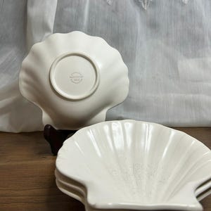 May include: Three off-white, shell-shaped dishes stacked together. The top dish faces upwards, while the base of another dish reveals a circular stamp with text. The dishes are on a wooden surface. The shell design is visible.