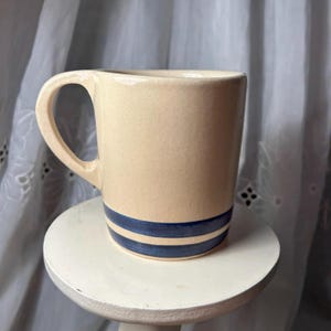 Könnte beinhalten: Eine cremefarbene Keramiktasse mit rundem Henkel und zwei marineblauen Streifen am Boden. Die Tasse ist zylindrisch und steht auf einem kleinen, weißen, runden Sockel.