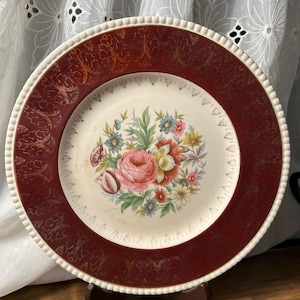 May include: A vintage decorative plate with a burgundy rim and a floral center. The plate features a white center with a bouquet of pink, blue, and yellow flowers. The rim has a gold filigree design and a white beaded edge.